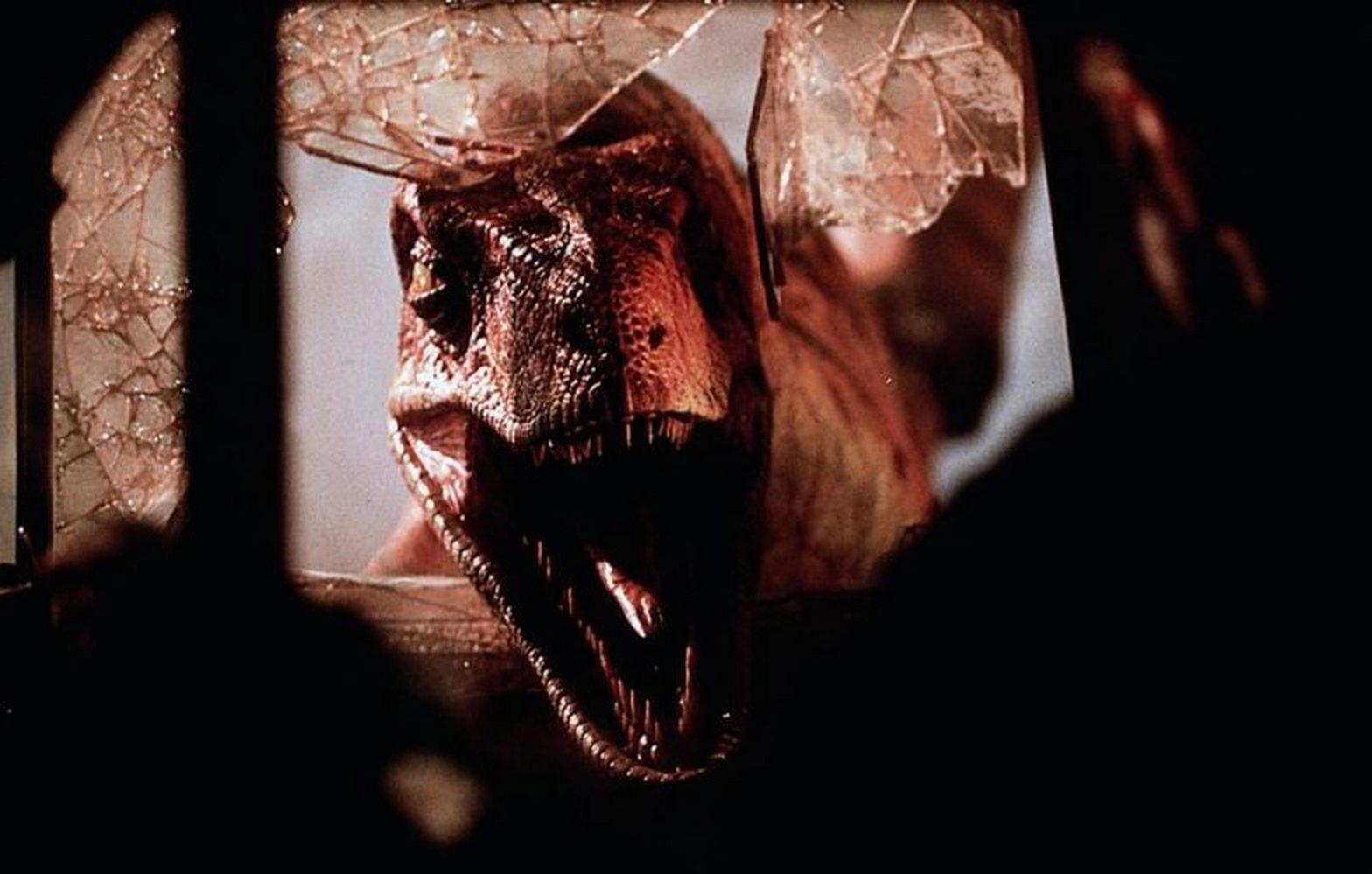 Nahaufnahme eines Dinosaurierkopfes, der durch eine zerbrochene Fensterscheibe bricht. Der Dinosaurier hat scharfe Zähne und einen bedrohlichen Ausdruck.