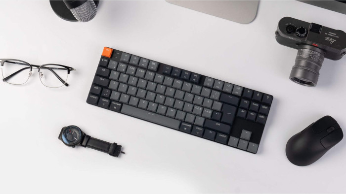 Auf einer weißen Oberfläche befinden sich eine Tastatur, eine Uhr, eine Brille, eine Kamera und eine Maus. Die Tastatur ist zentriert.