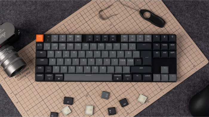 Mechanische Tastatur auf einer Schneidematte, Kamera und Maus daneben. Tastenkappen verstreut.