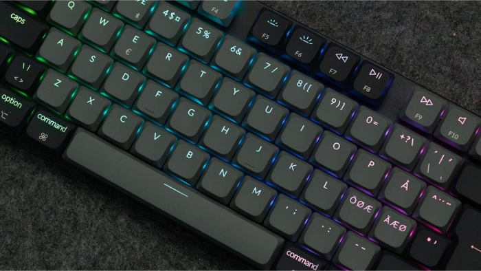 Nahaufnahme einer grauen mechanischen Tastatur. Tasten sind mit bunten Lichtern beleuchtet. Der Hintergrund ist eine strukturierte, dunkelgraue Oberfläche.