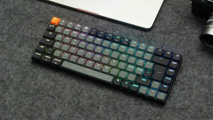 Eine grau-schwarze mechanische Tastatur mit RGB-Beleuchtung. Ein Laptop und eine Kamera befinden sich im Hintergrund auf einer grauen Oberfläche.