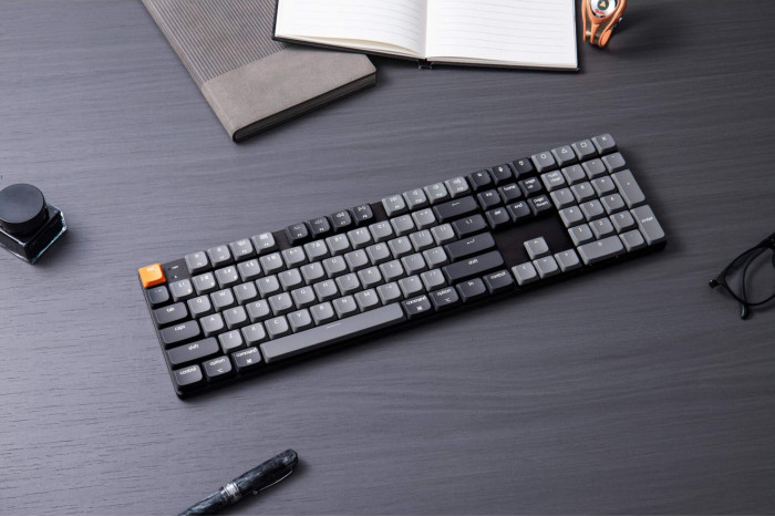 Eine Tastatur mit grauen und schwarzen Tasten auf einem dunklen Schreibtisch, mit Notizbuch, Stift, Tinte und Brille.