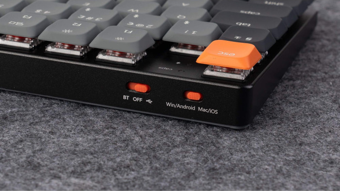 Nahaufnahme einer Tastatur. Graue Tasten mit weißen Buchstaben. Orangefarbene Escape-Taste. Schwarzer Rahmen. Zwei orangefarbene Schalter und Text darunter.