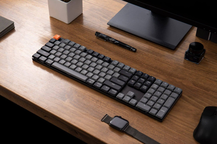 Eine Tastatur, ein Stift und eine Uhr liegen auf einem Holztisch. Ein Computermonitor und Zubehör befinden sich im Hintergrund. Die Tastatur hat schwarze und graue Tasten.