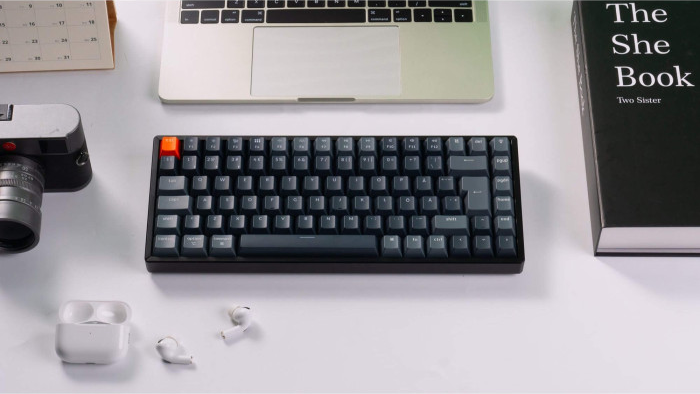 Eine Tastatur, ein Laptop, eine Kamera, ein Buch, ein Kalender und Ohrhörer liegen auf einer weißen Oberfläche. Die Tastatur hat eine orangefarbene Esc-Taste.