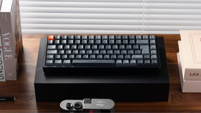 Eine schwarze Tastatur mit grauen Tasten, einer orangefarbenen Escape-Taste, auf einem schwarzen Ständer. Bücher und eine Kamera daneben.