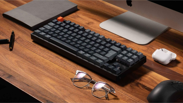 Ein Holztisch mit Tastatur, Brille, Stift, Notizbuch, Computer und Maus. Die Tastatur hat eine orangefarbene Escape-Taste.