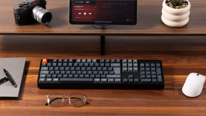 Ein Holztisch mit Tastatur, Maus, Brille und Stift. Ein Tablet zeigt Musik, eine Kamera und eine kleine Pflanze.