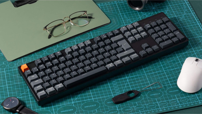 Eine schwarze Tastatur mit grauen Tasten und einer orangefarbenen Escape-Taste auf einer grünen Schneidematte, mit Brille, Maus und Uhr.