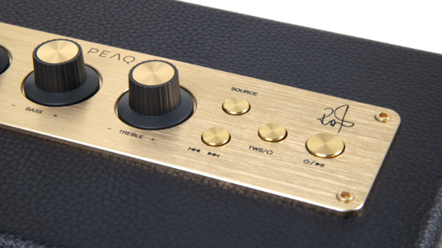 Zbliżenie panelu sterowania głośnika. Zawiera pokrętła, przyciski i podpis na złotej płycie. Czarne skórzane tło.