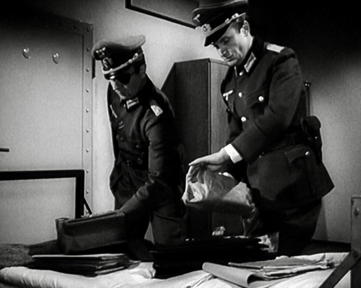 Zwei Männer in Uniform untersuchen in einem schwach beleuchteten Raum Dokumente und eine Tasche. Einer hält eine Tasche, der andere schaut sich einen Aktenkoffer an.