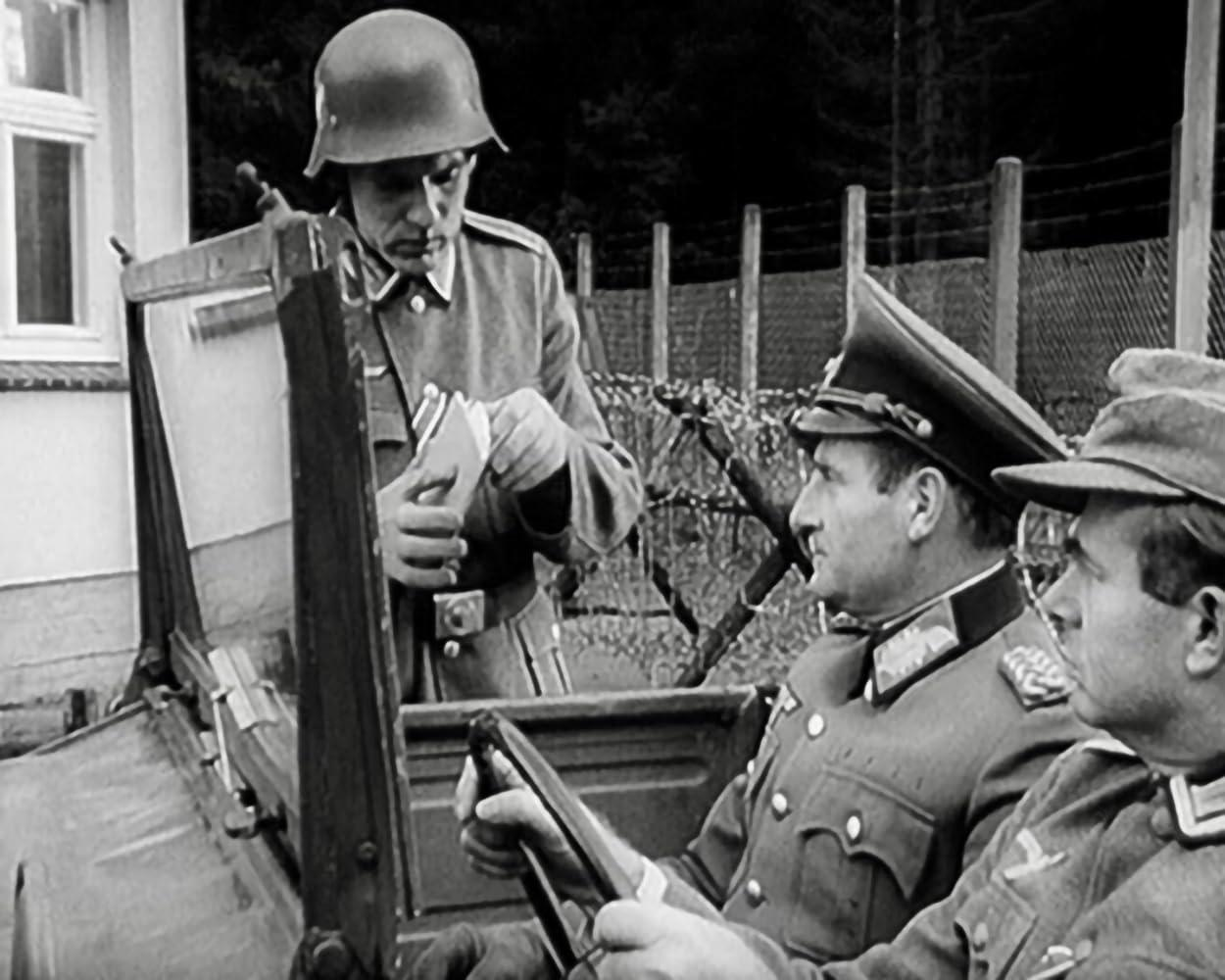 Soldaten in Uniform stehen in der Nähe eines Fahrzeugs. Ein Soldat mit Helm hält Papiere. Zwei Offiziere sitzen im Fahrzeug.
