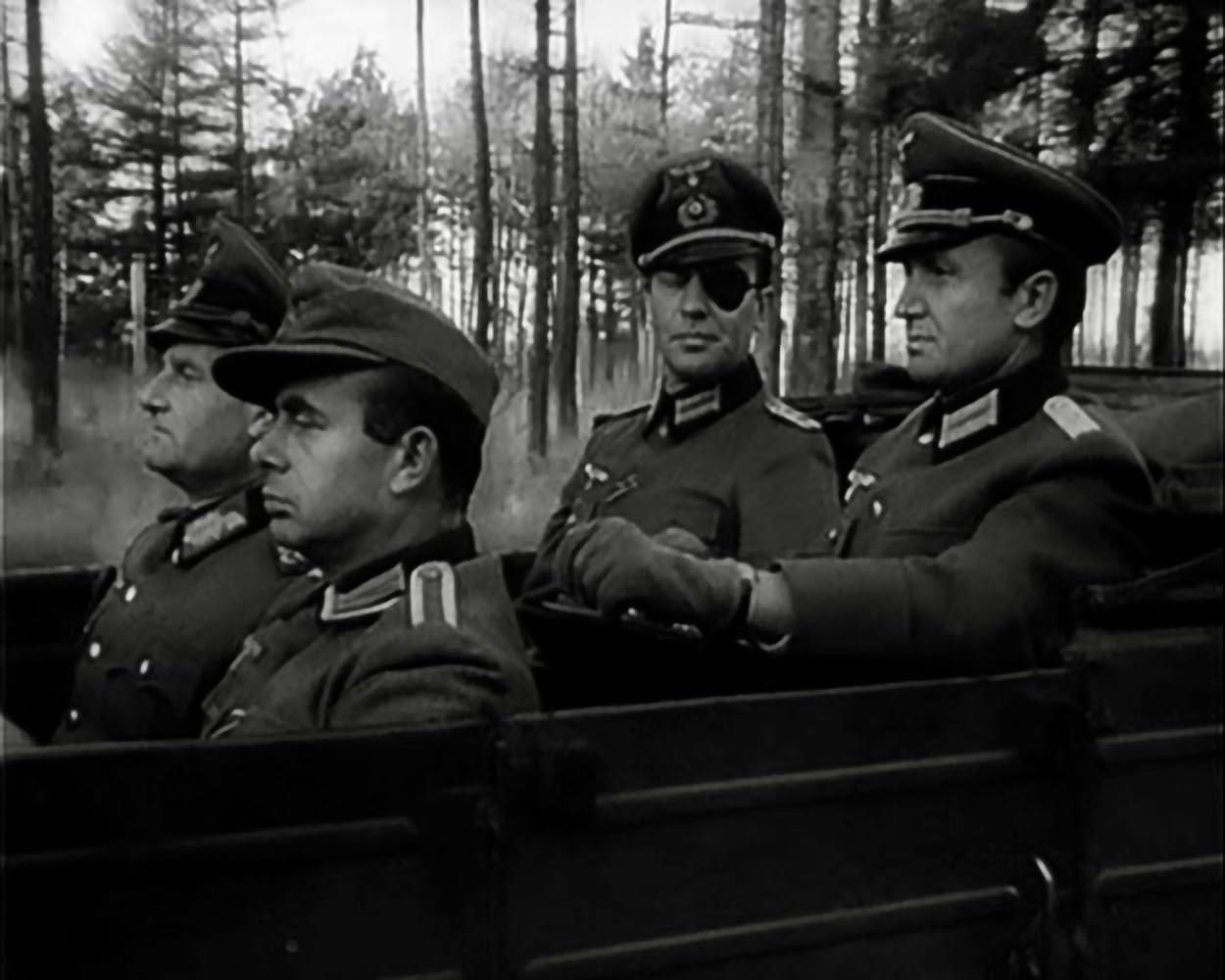 Vier Männer in Militäruniformen fahren in einem Fahrzeug. Einer trägt eine Augenklappe. Hohe Bäume sind im Hintergrund.