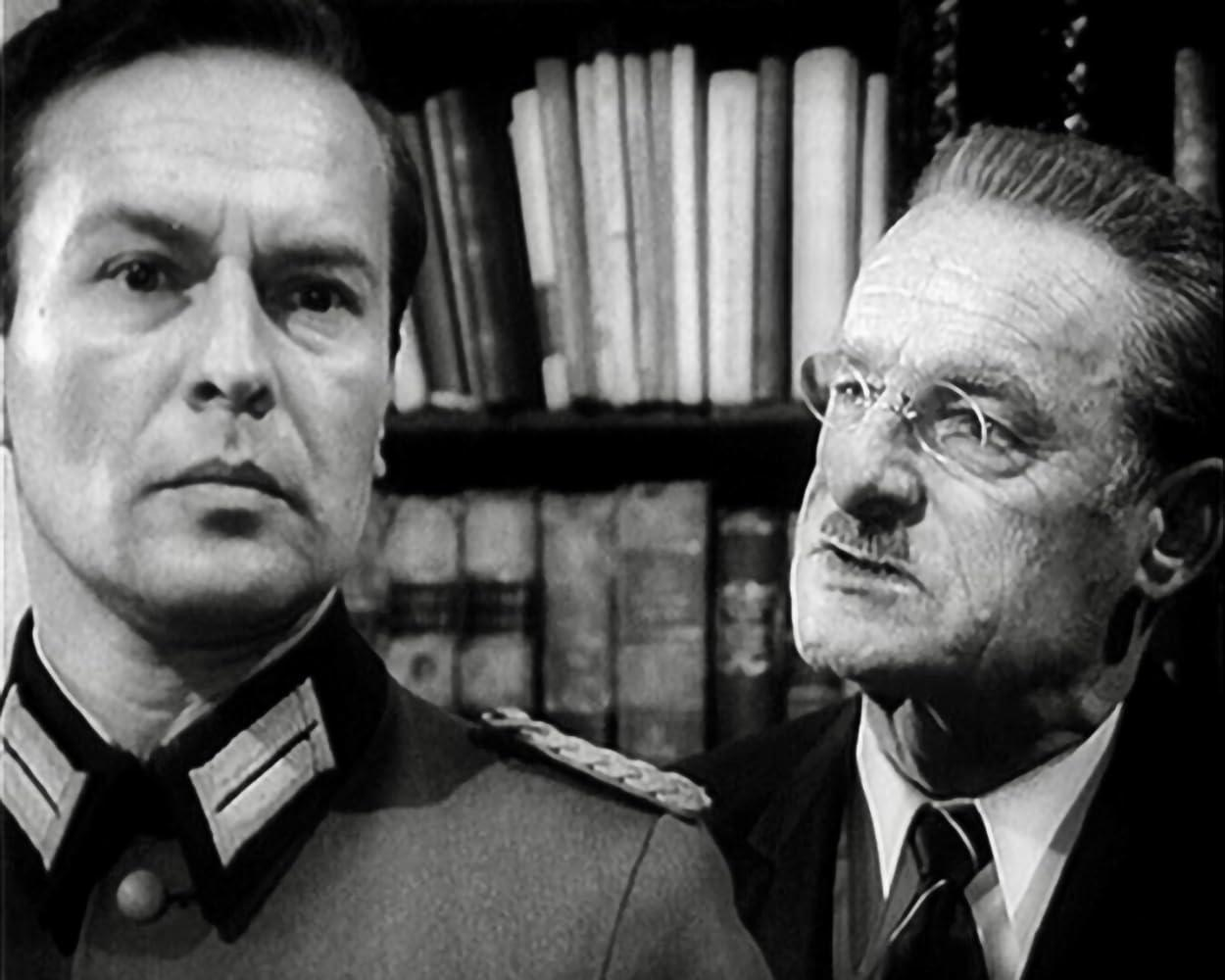Zwei Männer in Schwarzweiß. Einer in Uniform, der andere im Anzug mit Brille. Bücherregale im Hintergrund.