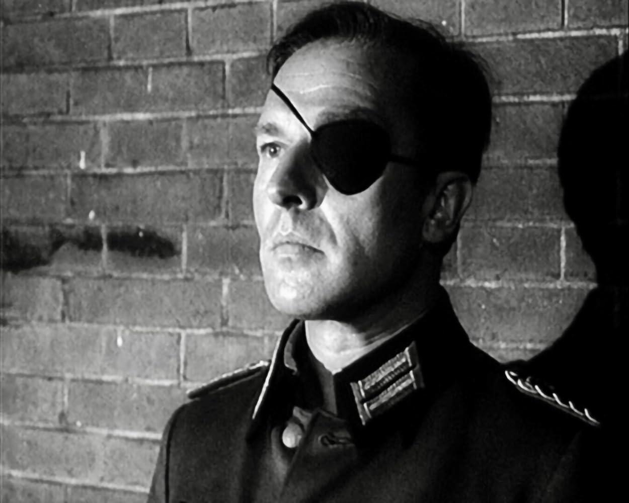 Ein Mann mit Augenklappe trägt eine Uniform. Er steht vor einer Ziegelmauer. Das Bild ist in Schwarzweiß gehalten.