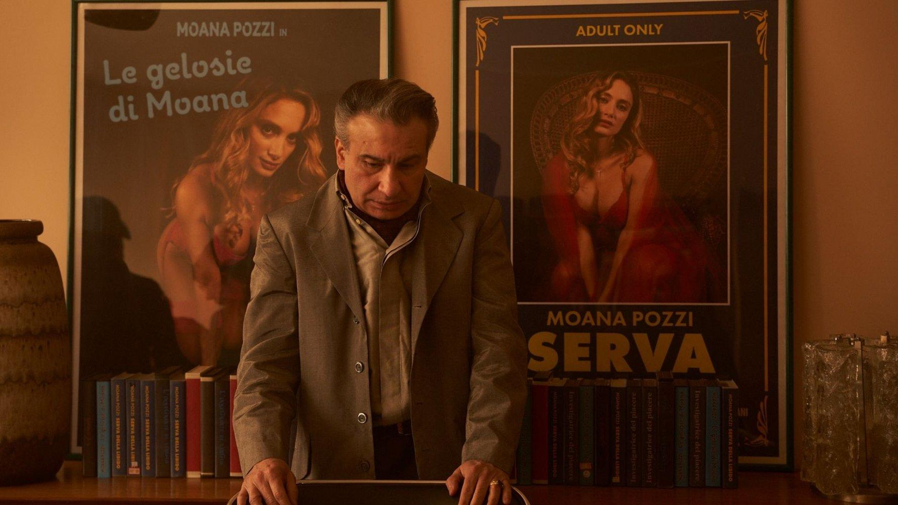 Ein Mann im Anzug steht vor zwei Postern und einem Bücherregal. Die Poster zeigen eine Frau. Der Mann blickt nach unten.