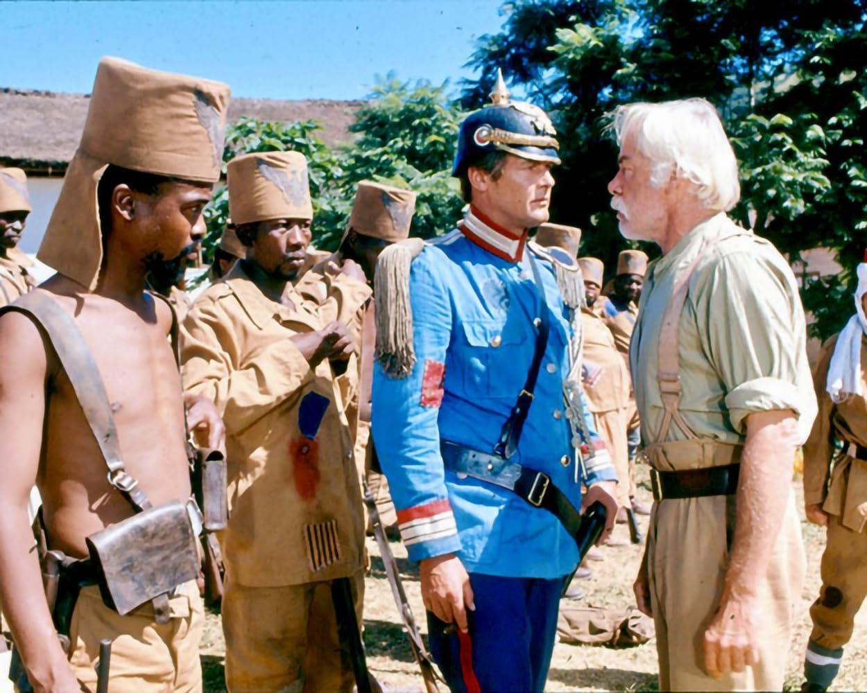 Männer in verschiedenen Uniformen stehen draußen. Einer in Blau steht einem Mann in Khaki gegenüber. Andere tragen beige Uniformen und Hüte. Grüne Bäume im Hintergrund.