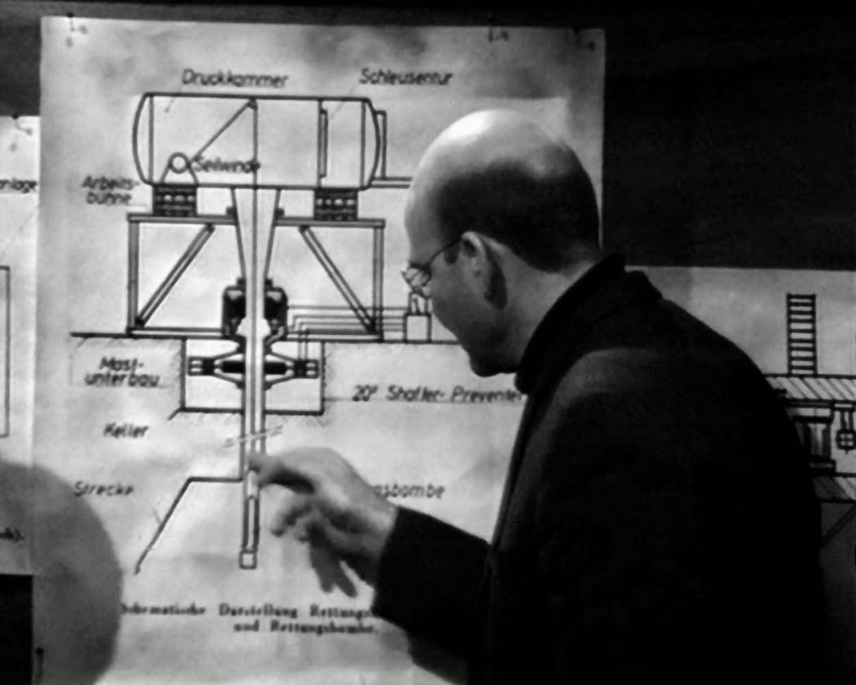 Ein Mann im dunklen Anzug zeigt auf eine Zeichnung einer Industriestruktur mit deutschen Beschriftungen. Das Diagramm enthält einen Tank und andere Teile.