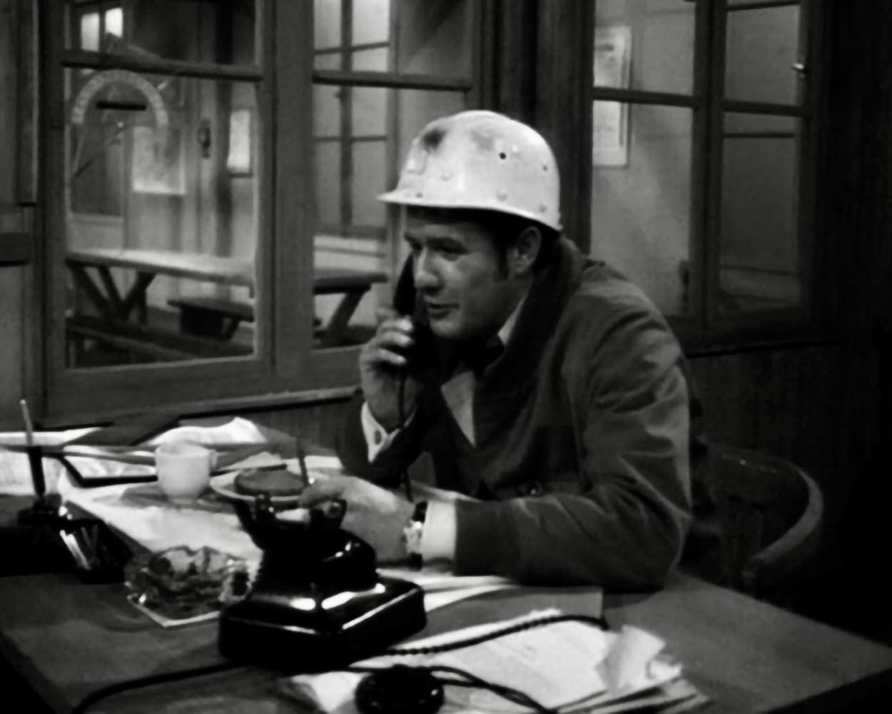 Ein Mann mit Schutzhelm telefoniert in einem Büro. Auf dem Schreibtisch befinden sich ein schwarzes Telefon, Papiere und eine Tasse.