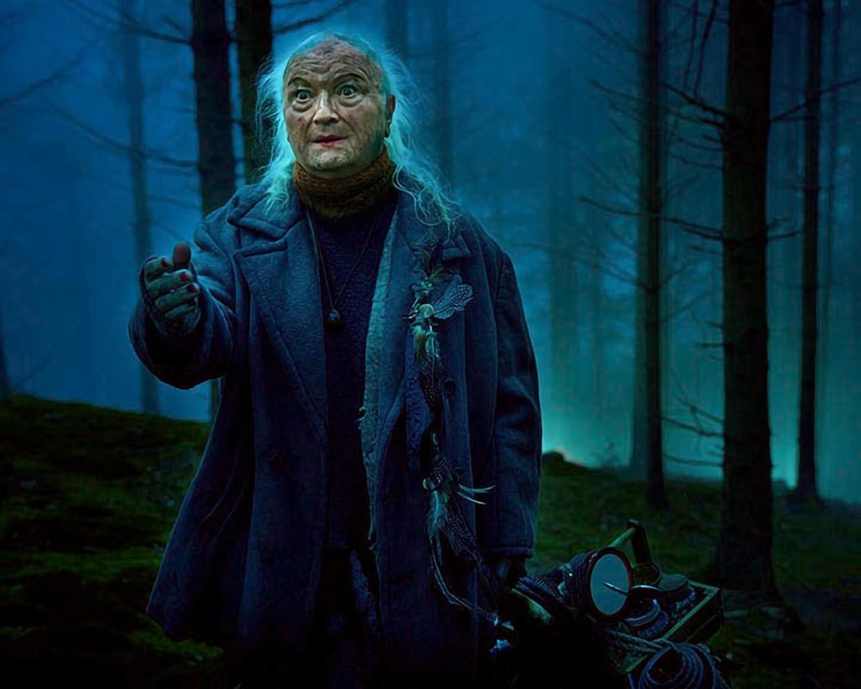 Eine ältere Frau in einem blauen Mantel gestikuliert in einem nebligen Wald. Sie hält eine Holzkiste und ist von Bäumen umgeben.