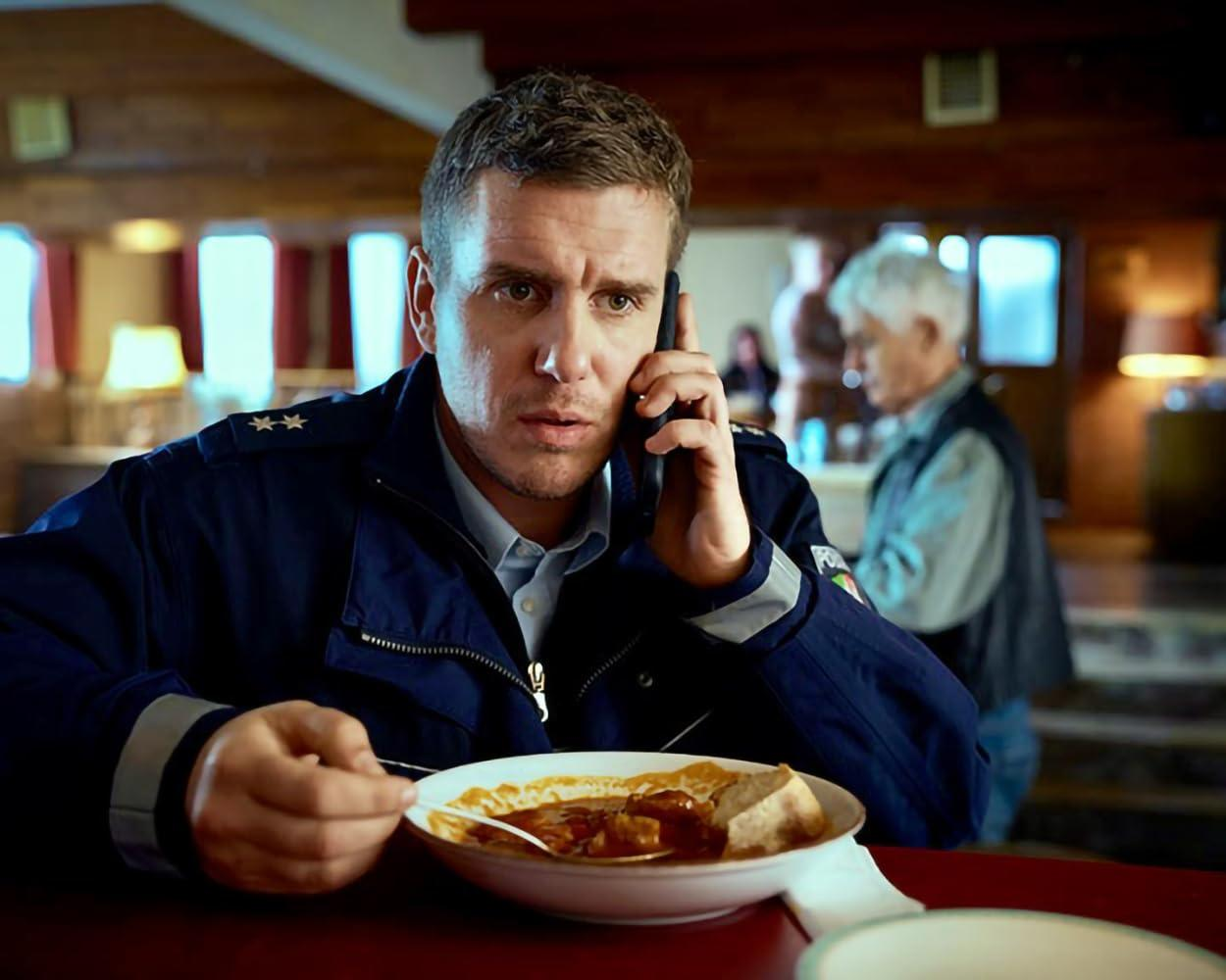 Ein Mann in Polizeiuniform telefoniert beim Essen eines Eintopfs. Im Hintergrund ist ein älterer Mann zu sehen. Die Szene spielt drinnen.