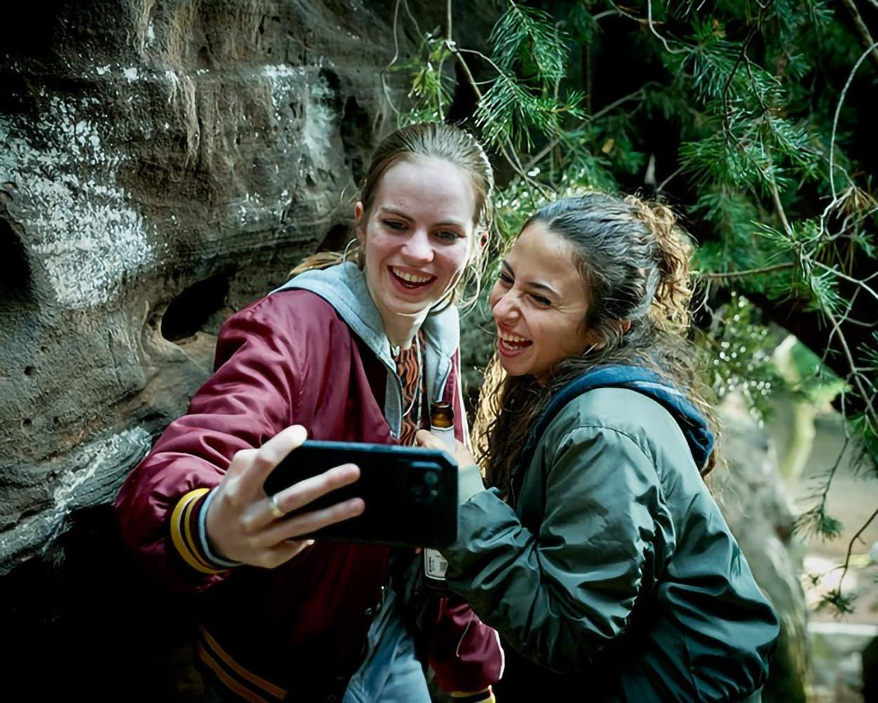 Zwei lächelnde Frauen machen ein Selfie mit einem Telefon. Eine trägt eine rote Jacke, die andere eine grüne Jacke, beide in der Nähe von Bäumen und einem Felsen.