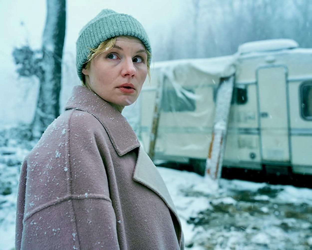 Eine Frau in einem rosa Mantel und einer grünen Mütze blickt in einer verschneiten Landschaft zurück. Im Hintergrund ist ein Wohnwagen zu sehen.