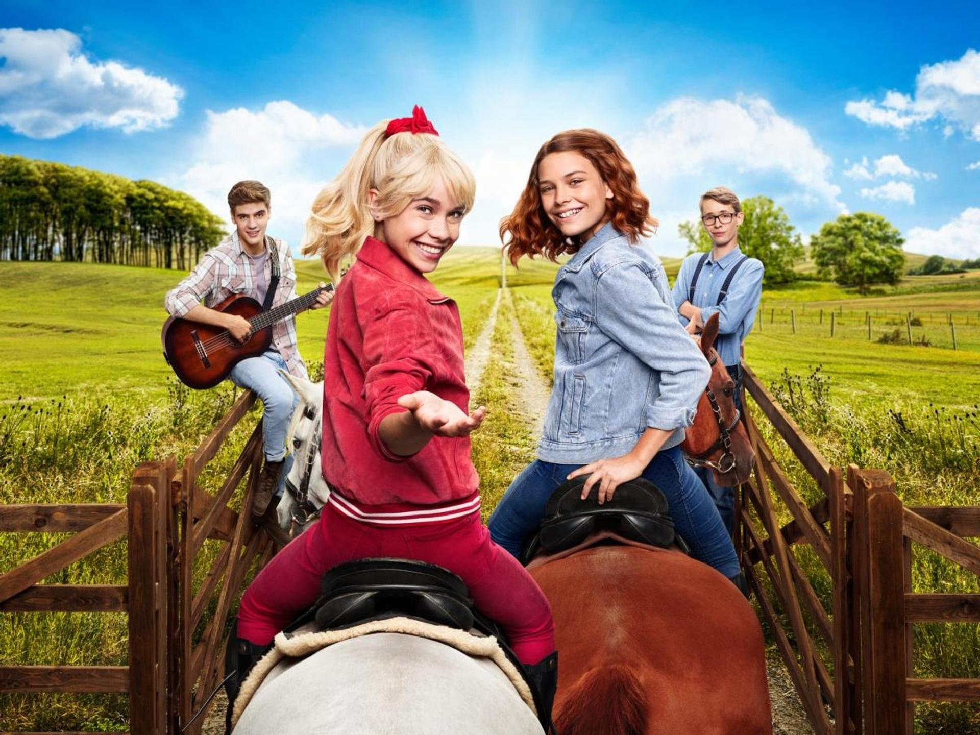Vier junge Leute auf Pferden und ein Gitarrist auf einem Feld mit blauem Himmel.