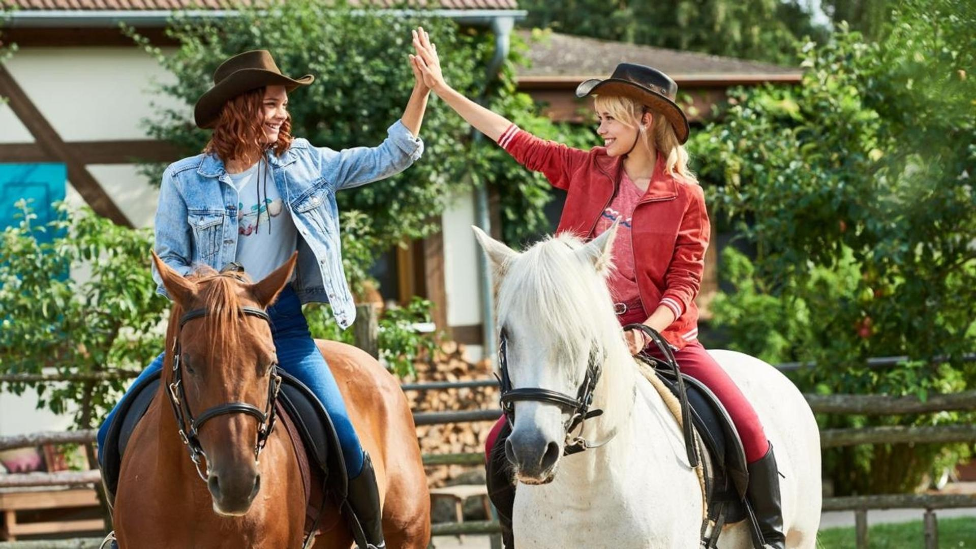 Zwei Frauen auf Pferden geben sich High-Five. Eine reitet auf einem braunen Pferd und trägt eine Jeansjacke.