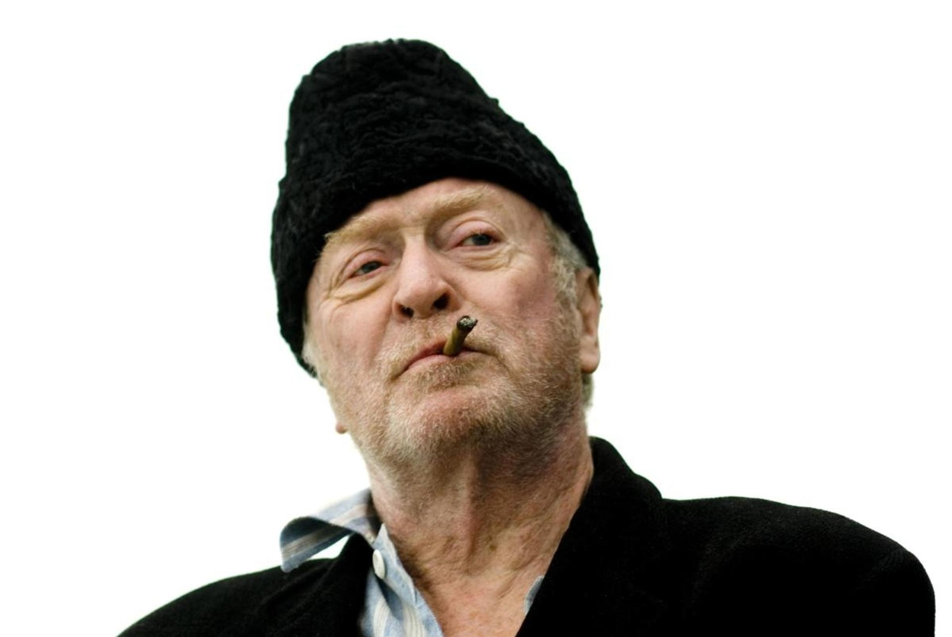 Ein älterer Mann mit schwarzem Pelzhut, der eine Zigarre raucht. Er hat einen weißen Hintergrund.