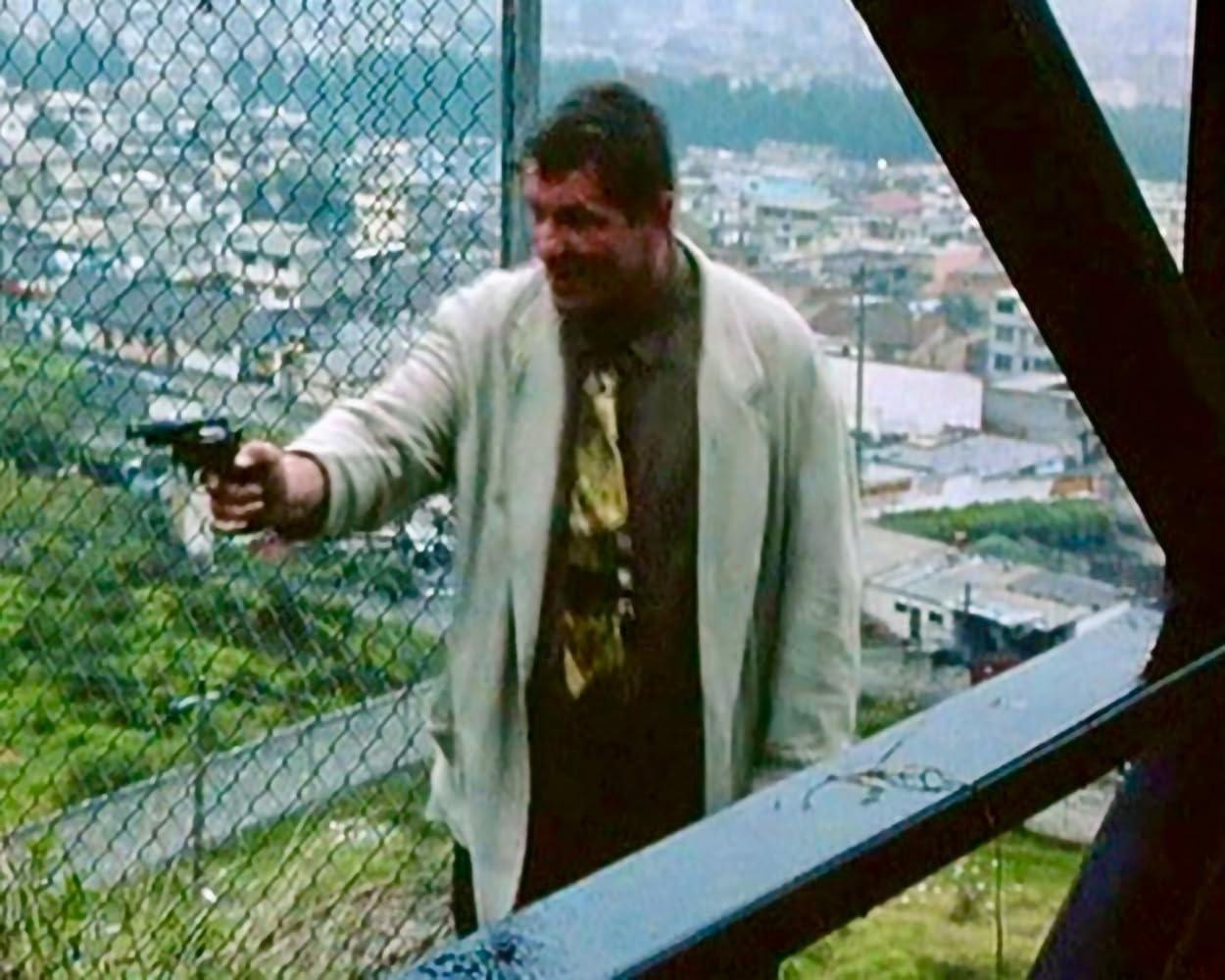 Ein Mann in einer Jacke hält eine Waffe. Er steht in der Nähe eines Zauns, im Hintergrund eine Stadtlandschaft.