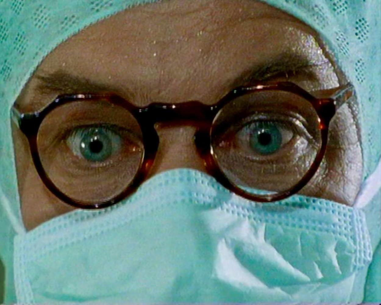 Nahaufnahme von Augen mit Brille, OP-Haube und Maske. Blaue Augen sind sichtbar. Die Szene deutet auf eine medizinische Umgebung hin.