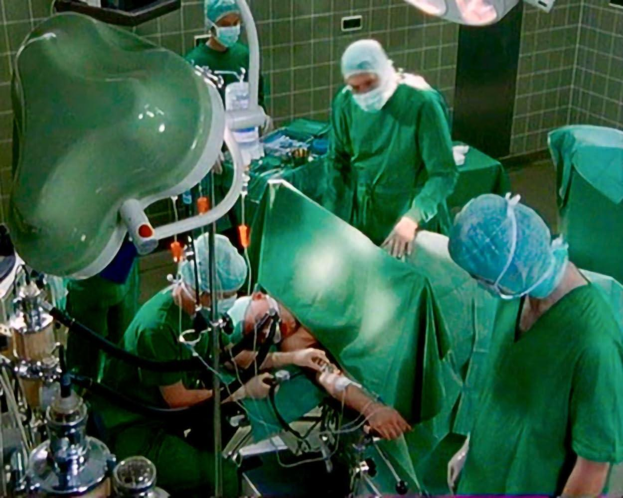 Operationssaal mit Chirurgen in grüner Kleidung um einen Patienten. Medizinische Geräte und Beleuchtung sind vorhanden. Fokus auf den Arm des Patienten.