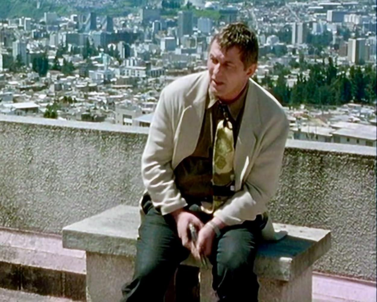 Ein Mann in einer hellen Jacke sitzt auf einer Bank mit Blick auf eine Stadt. Er trägt eine Krawatte und dunkle Hosen. Die Stadt ist im Hintergrund.