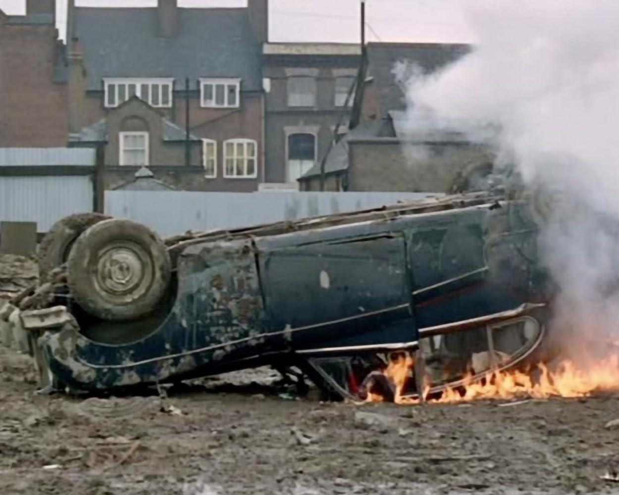 Ein Auto liegt auf dem Dach und brennt. Rauch steigt auf. Gebäude im Hintergrund. Die Szene zeigt einen Unfall.