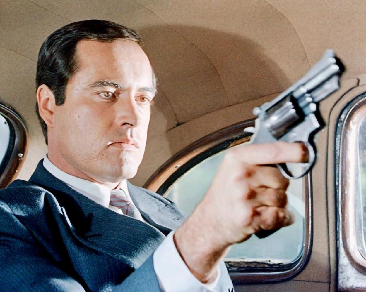 Ein Mann im Anzug zielt mit einer Waffe. Er befindet sich in einem Auto. Der Hintergrund zeigt das Innere des Autos.