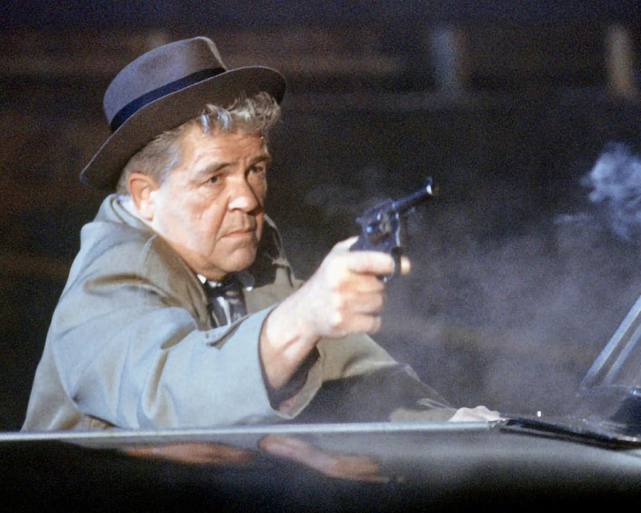 Ein älterer Mann mit Hut zielt mit einer Waffe, Rauch quillt hervor. Er trägt einen Mantel und ist in der Nähe eines Autos.