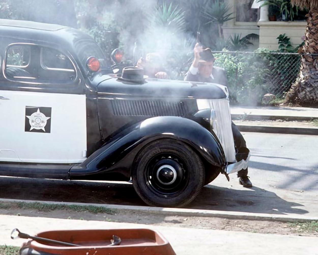 Schwarzes und weißes Polizeiauto mit zwei Männern. Ein Mann hält eine Waffe, Rauch steigt aus der Motorhaube. Roter Wagen im Vordergrund.