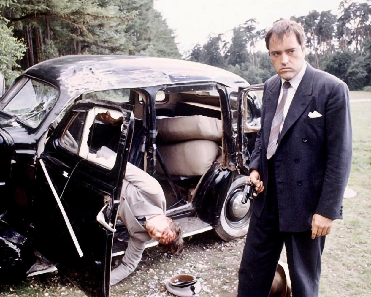 Ein Mann im Anzug steht neben einem beschädigten schwarzen Auto. Ein anderer Mann liegt im Inneren mit blutigem Kopf. Eine Pistole ist in der Hand des stehenden Mannes.