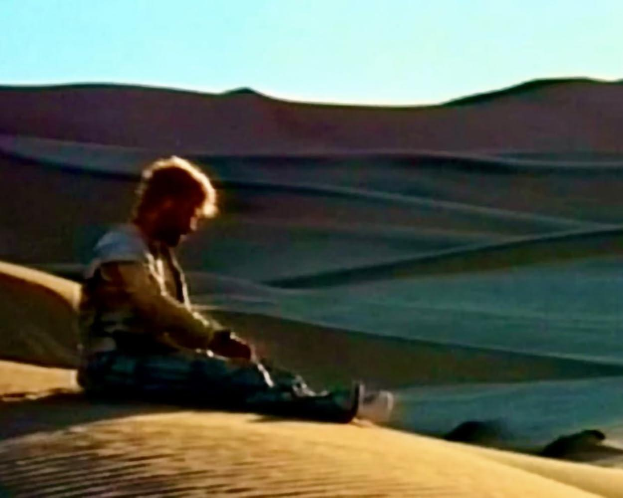 Ein Mann sitzt auf einer Sanddüne. Er trägt eine Weste und Hose. Der Hintergrund ist eine Wüste mit Dünen unter blauem Himmel.