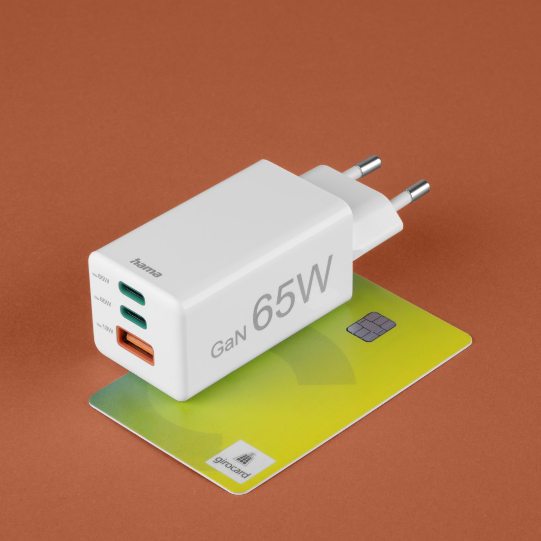 Ein weißes Hama GaN 65W Ladegerät liegt auf einer Girocard. Das Ladegerät befindet sich über einer gelb-grünen Kreditkarte, alles auf orangefarbenem Hintergrund.