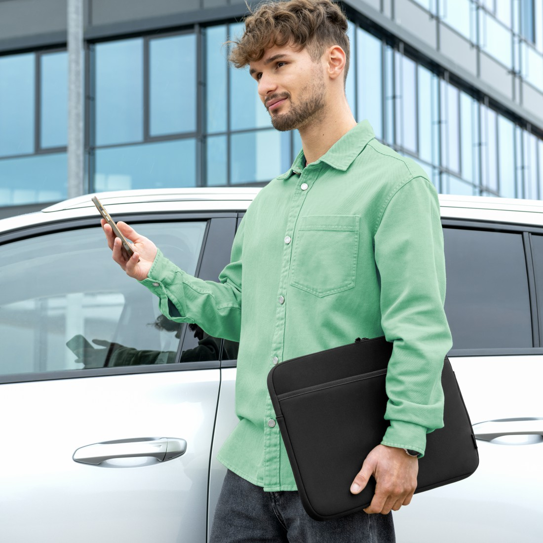 Mann in grünem Hemd hält Handy und schwarze Laptoptasche in der Nähe eines weißen Autos, im Hintergrund ein modernes Gebäude.