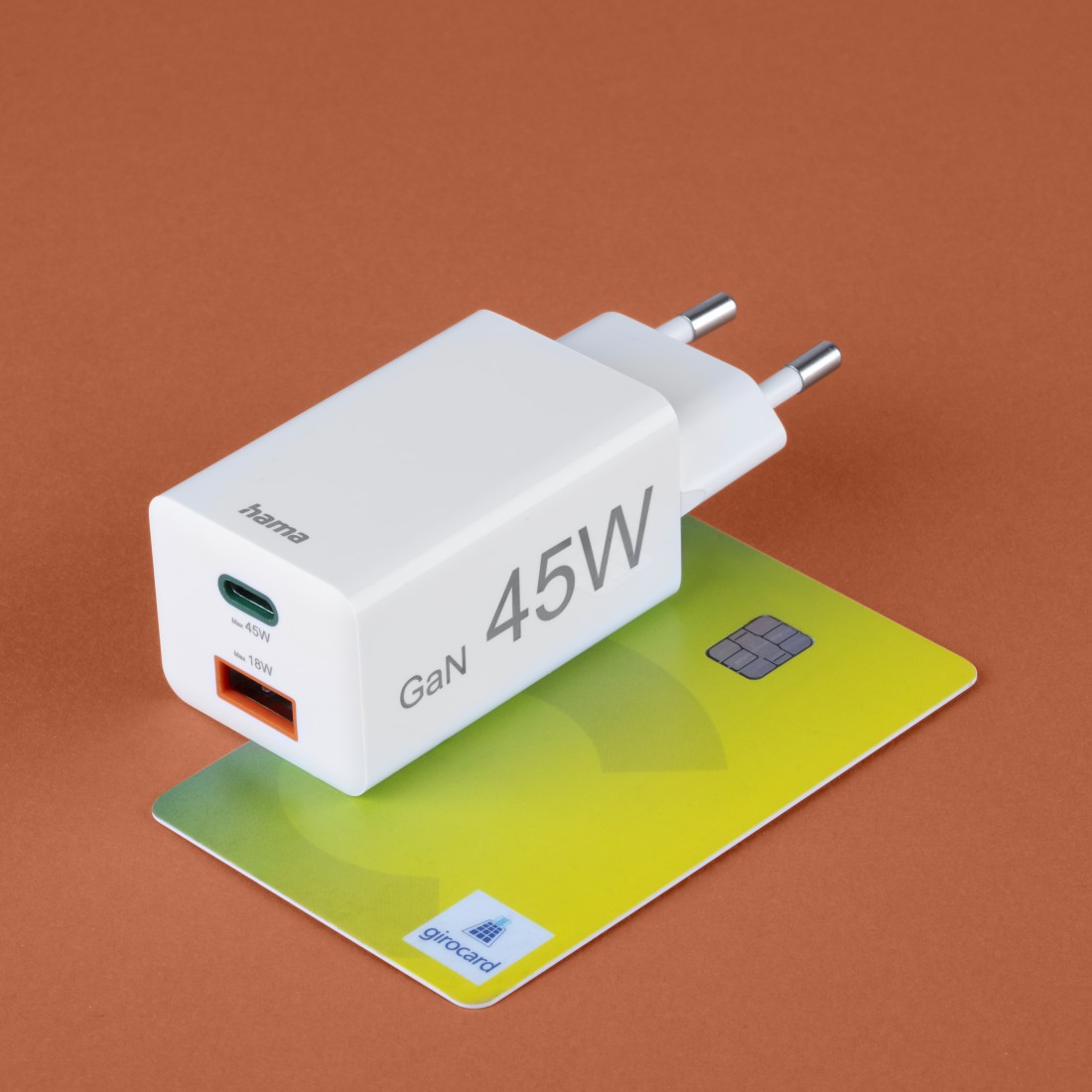 Ein weißes Hama 45W-Ladegerät liegt auf einer grün-gelben Girocard. Der Hintergrund ist orange.