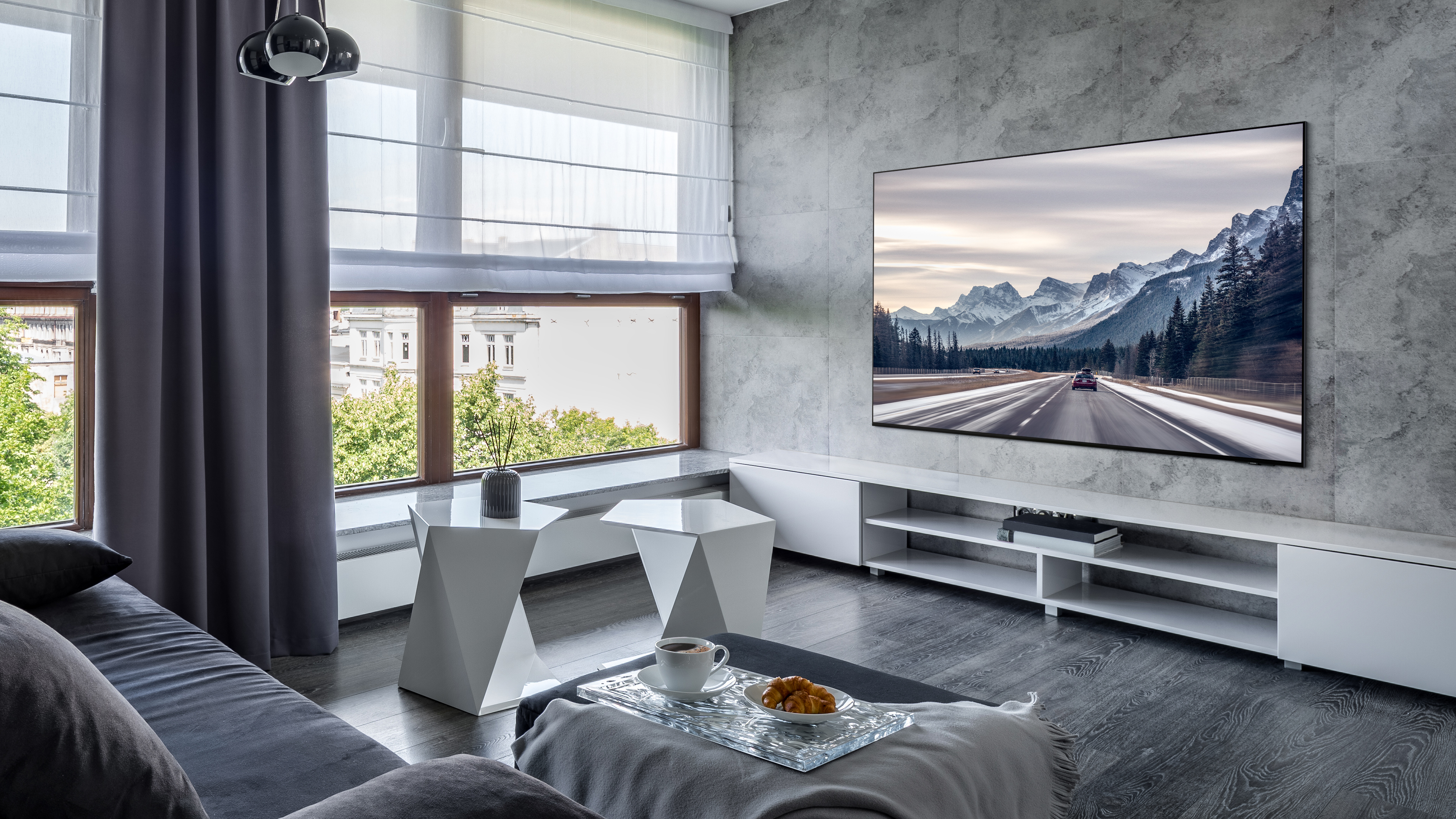 Un soggiorno moderno con una grande TV che mostra una scena di strada di montagna. C'è un divano, tavoli bianchi e una vista sulla finestra.