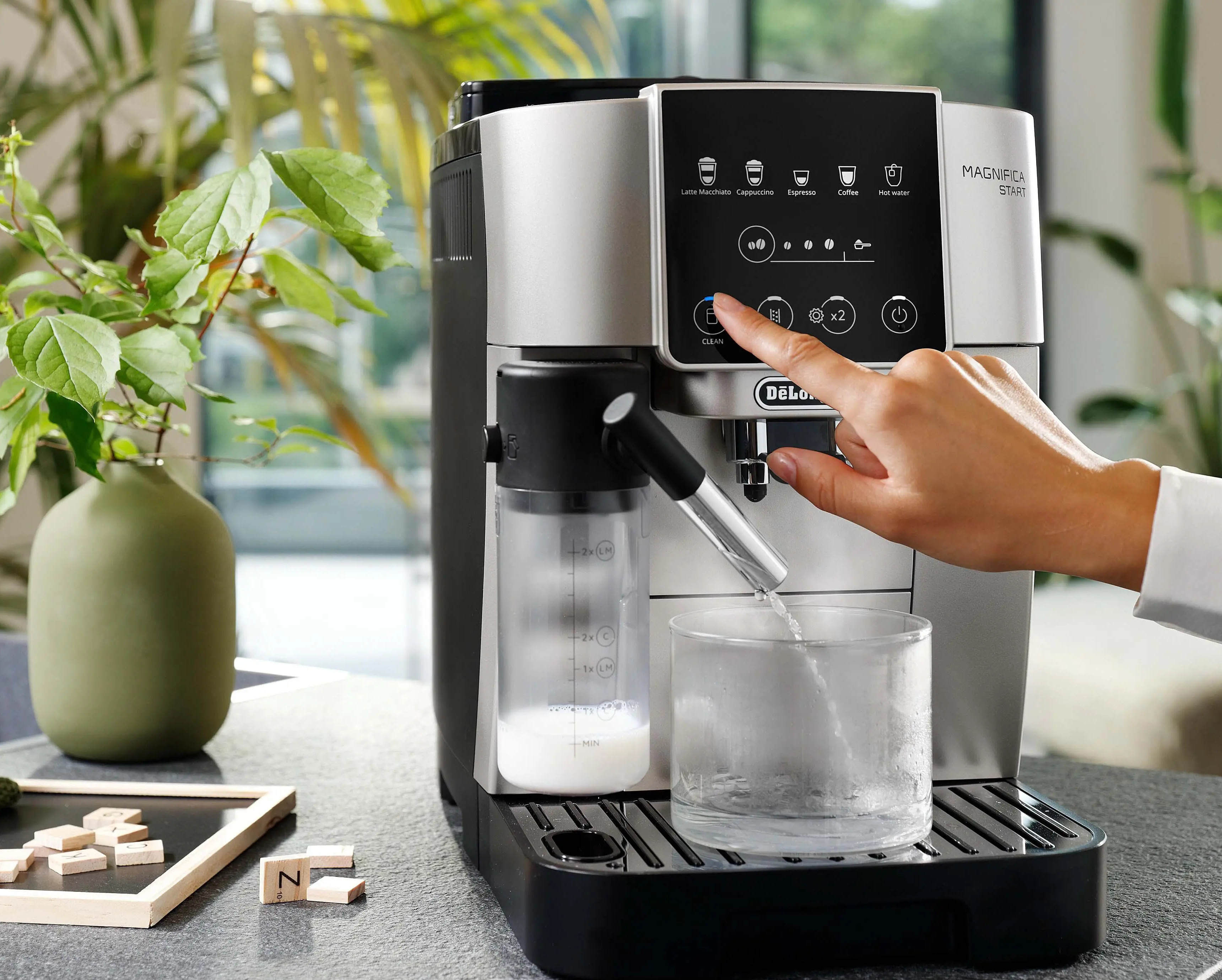 Srebrno-czarny ekspres do kawy. Ręka naciska przycisk. Szklanka jest napełniana wodą. Zielona roślina w tle.