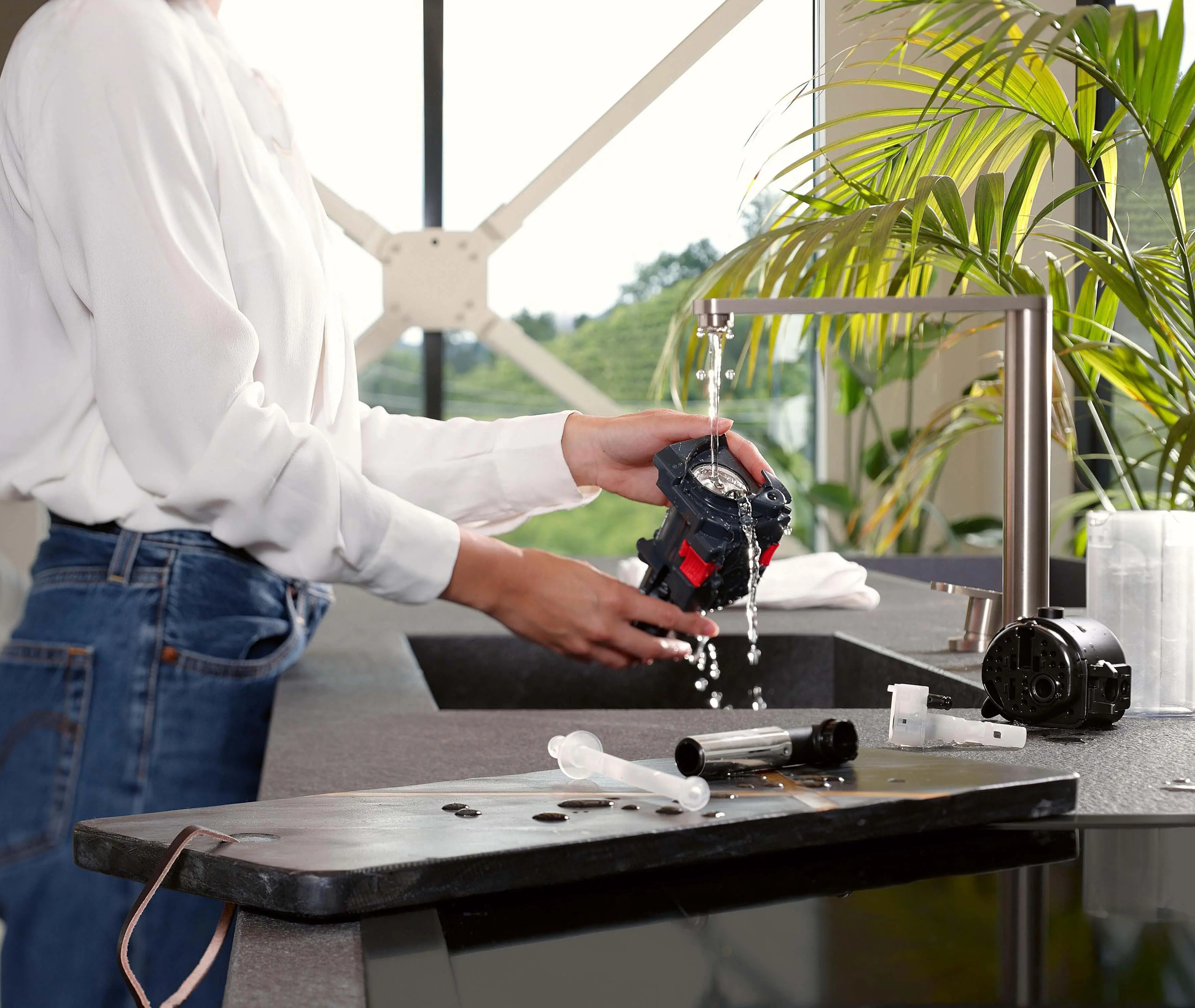 Osoba myje część ekspresu do kawy pod kranem. Inne części i roślina są na blacie.