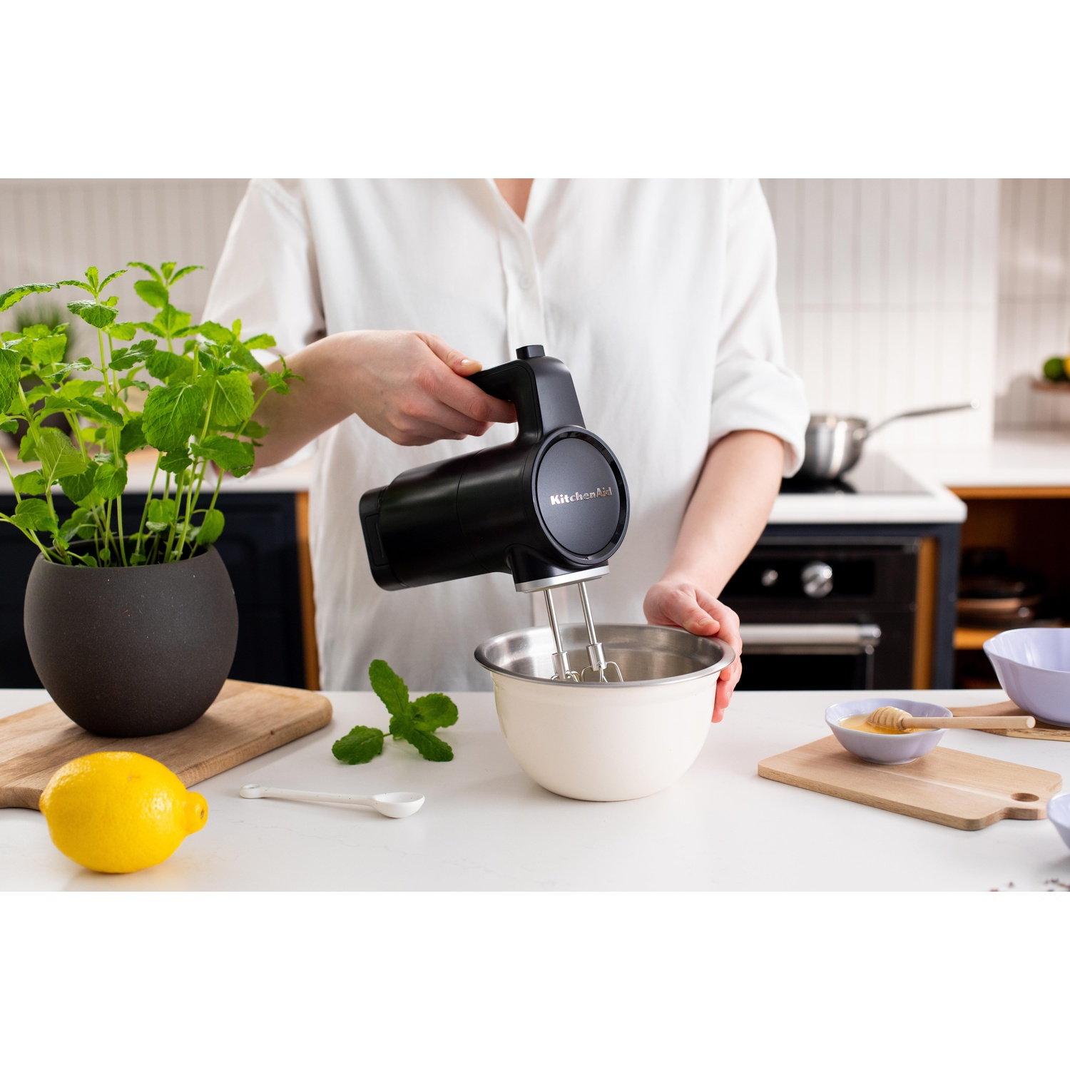 Persona usa un frullatore a mano KitchenAid nero in cucina. Ingredienti: limone, menta e ciotola. Lo sfondo include un fornello.