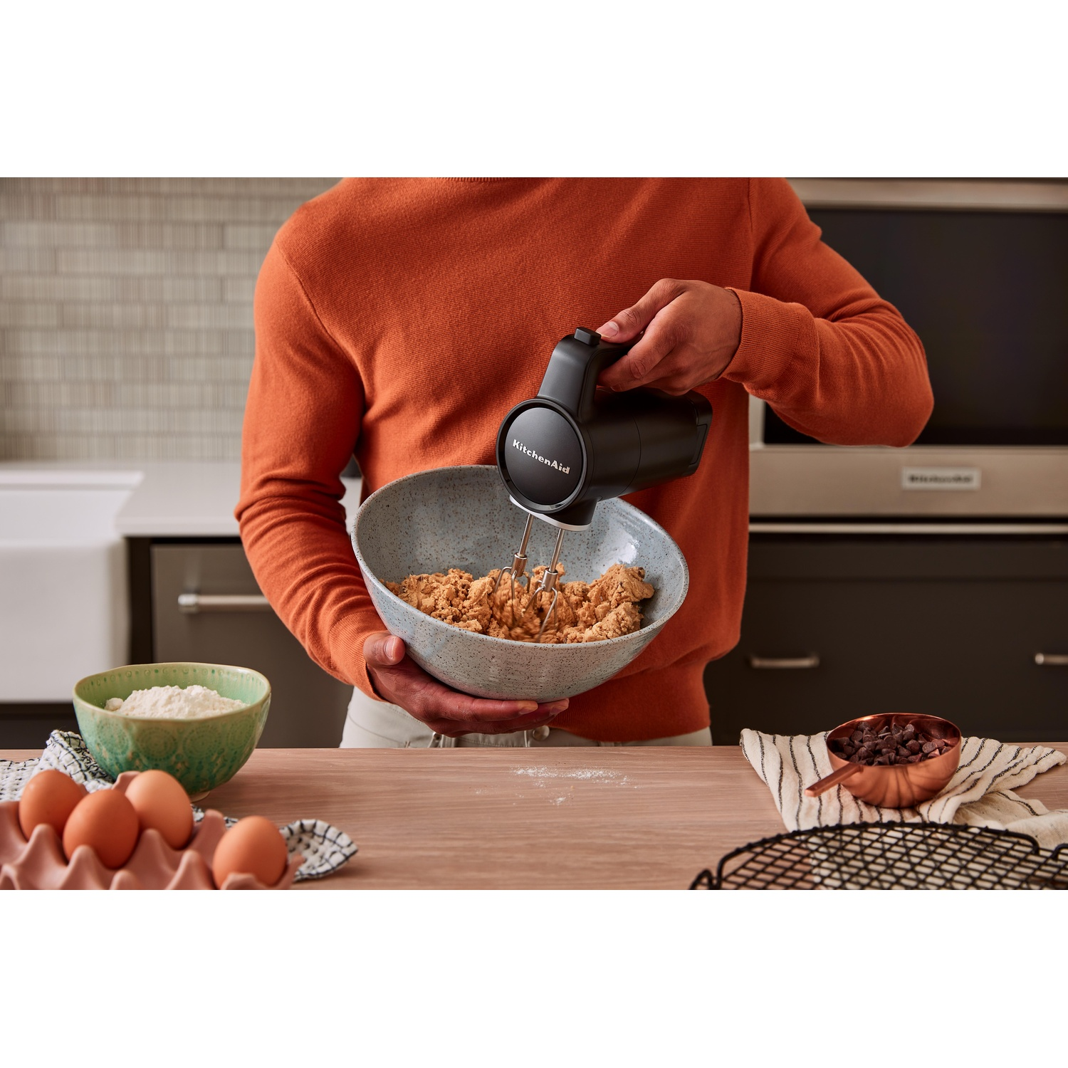 Persona che mescola gli ingredienti in una ciotola con un frullatore elettrico. Uova, farina e gocce di cioccolato sul tavolo. Sfondo della cucina.