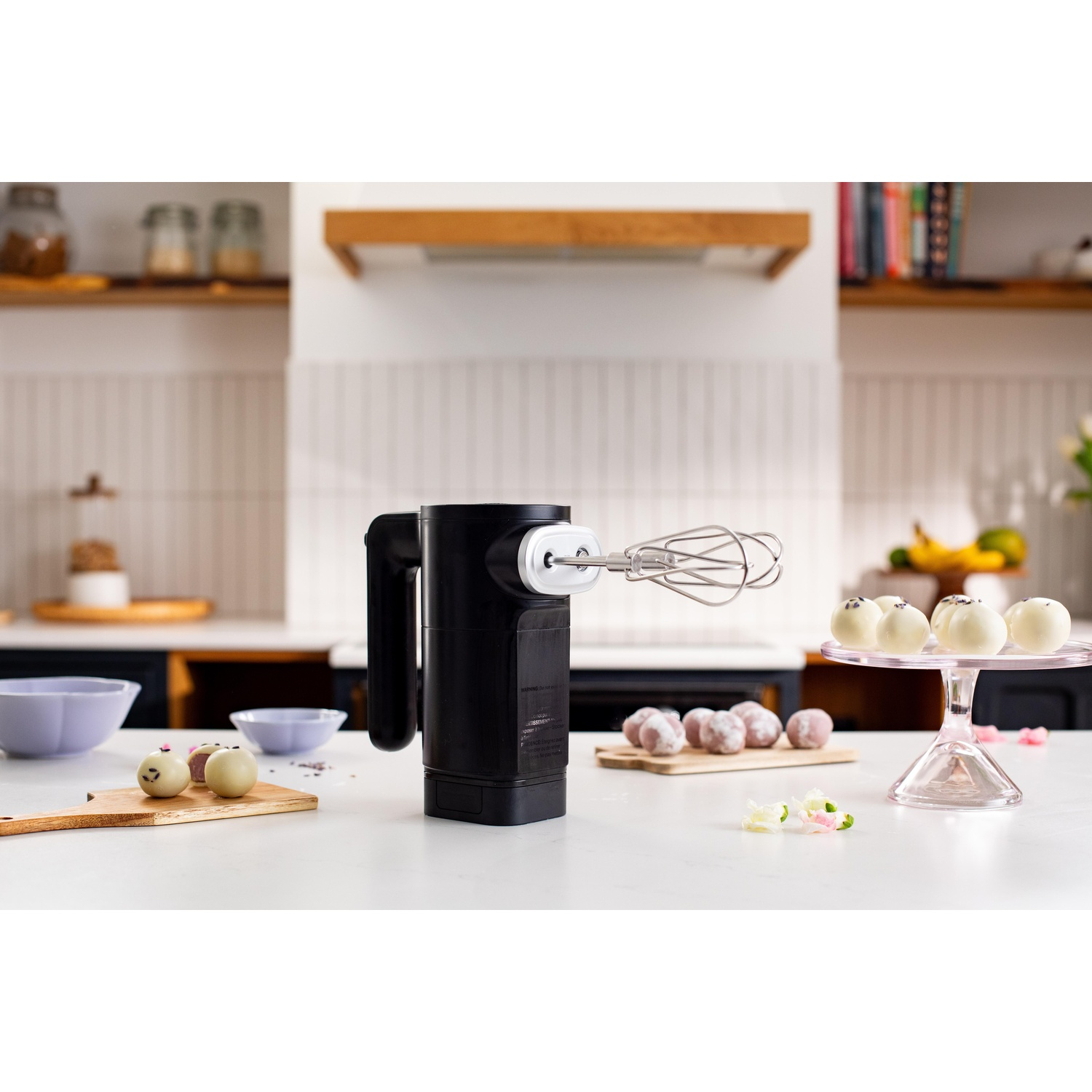 Un frullatore a mano nero con accessorio frusta è su un bancone bianco. Ci sono prodotti da forno e ciotole. Cucina sullo sfondo.