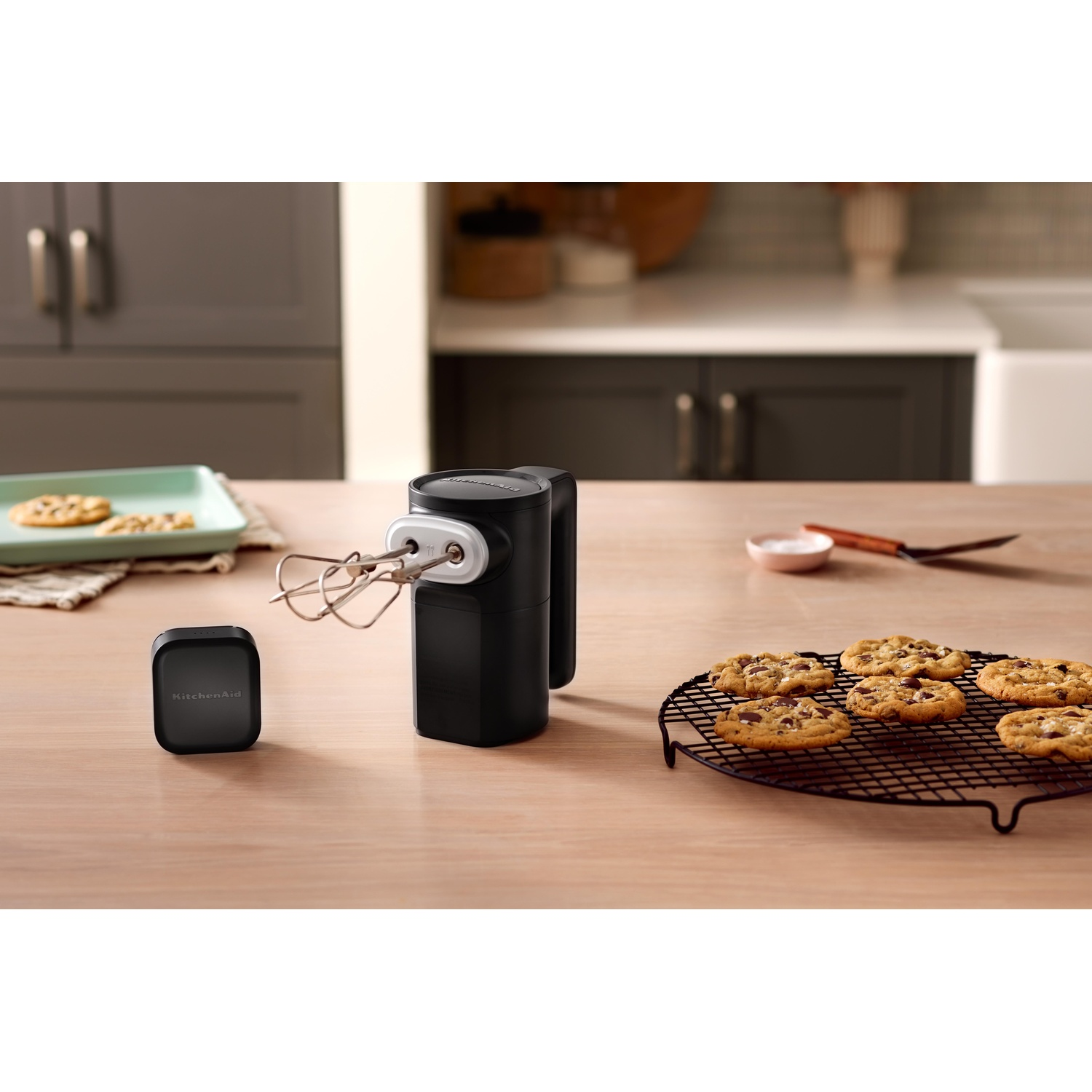 Un frullatore a mano KitchenAid nero con fruste, biscotti e una ciotola su un tavolo di legno in cucina.